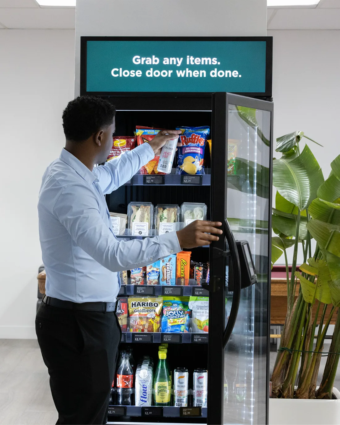 A person opening the glass door of a smart vending machine in a workplace break area and taking a snack from the shelf
