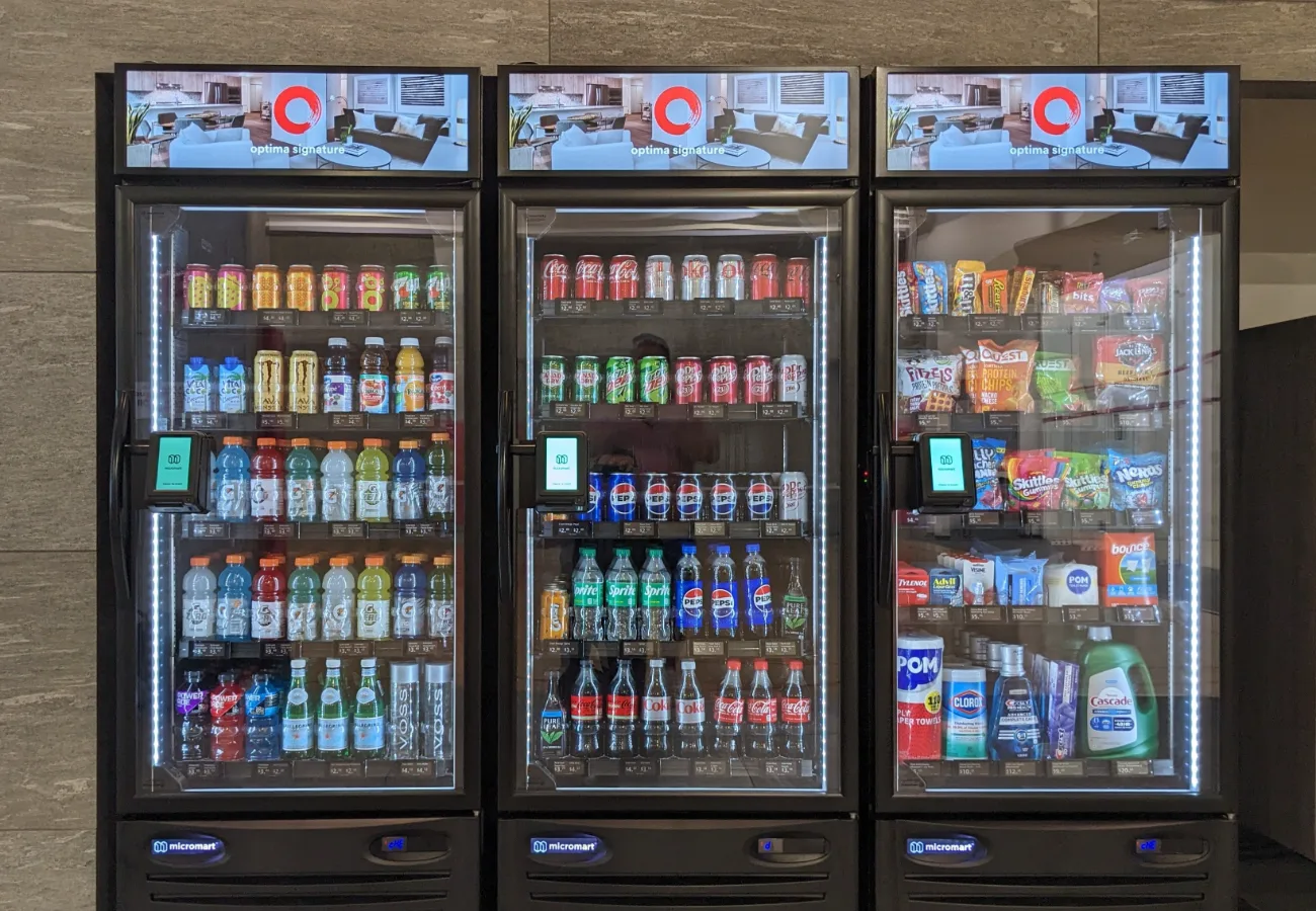 Three smart vending cabinets in a building lobby, each with a lit digital display above it carrying the property's own branding