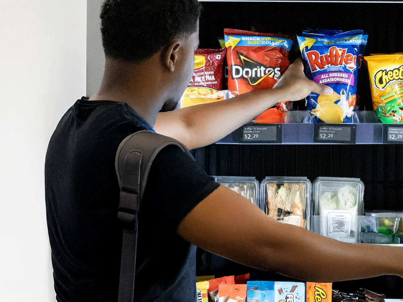 A shopper reaching into an open smart vending machine and lifting a bag of chips from a fully stocked shelf